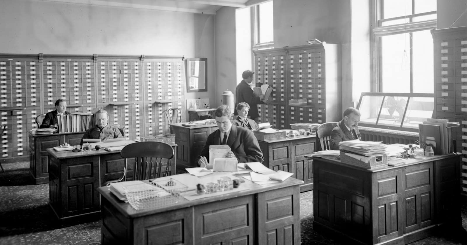 An early twentieth-century office, clerks at wooden desks among filing cabinets