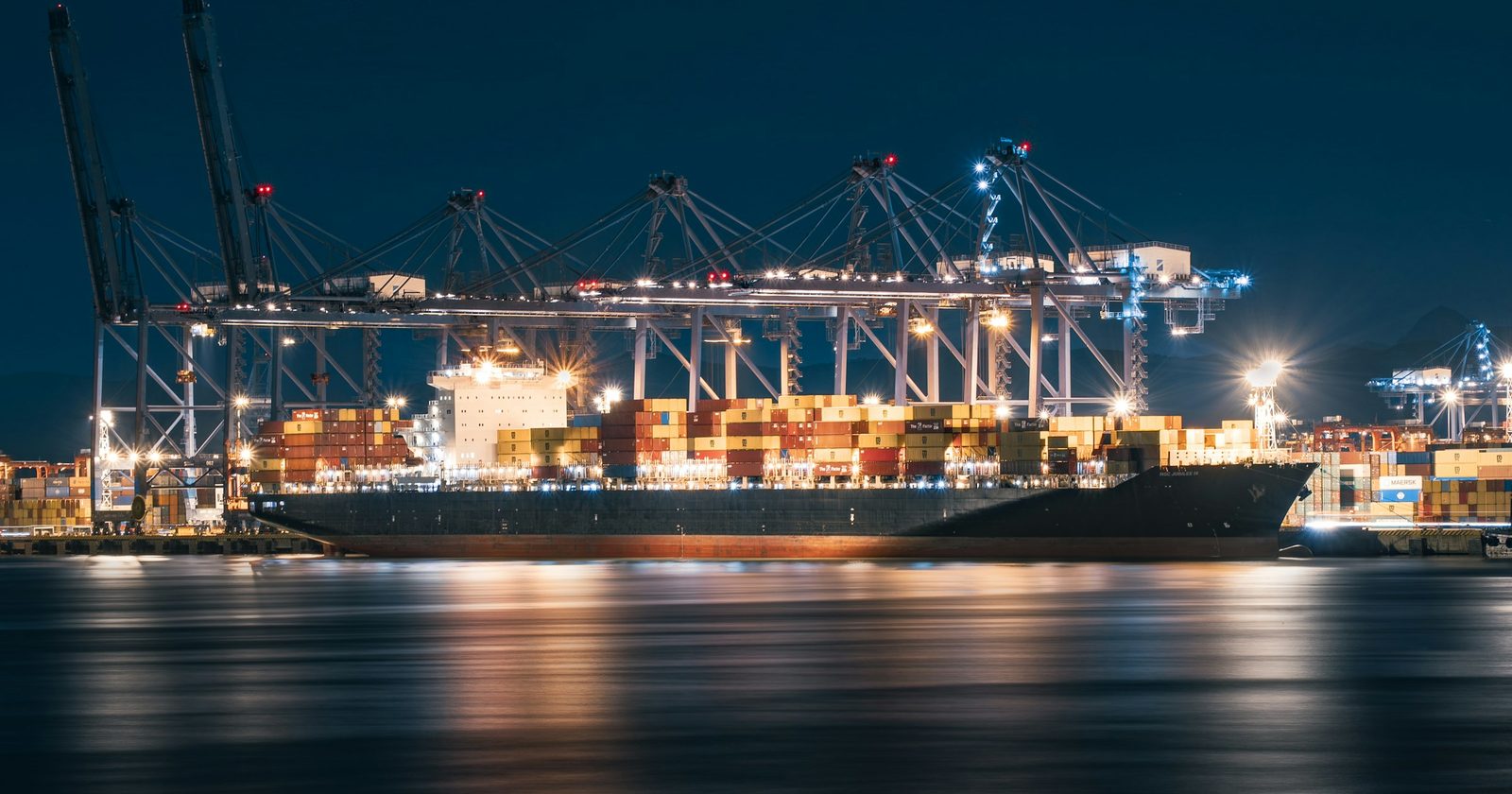 A cargo ship alongside a working port at night, cranes lit, the clock on its proceeds already running