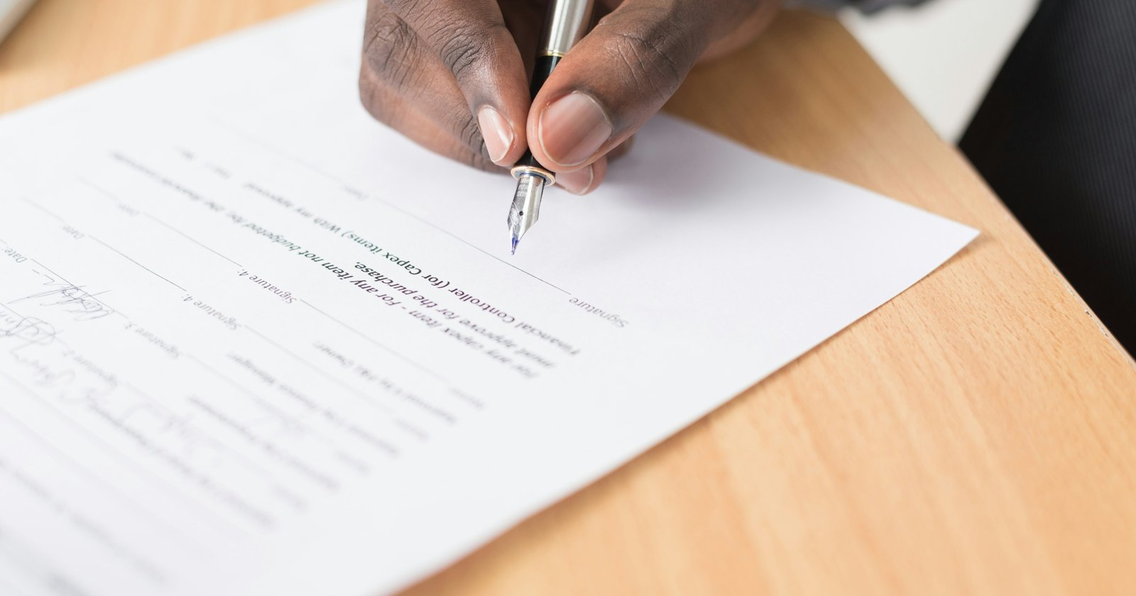 A hand signing a printed document with a fountain pen on a wooden desk