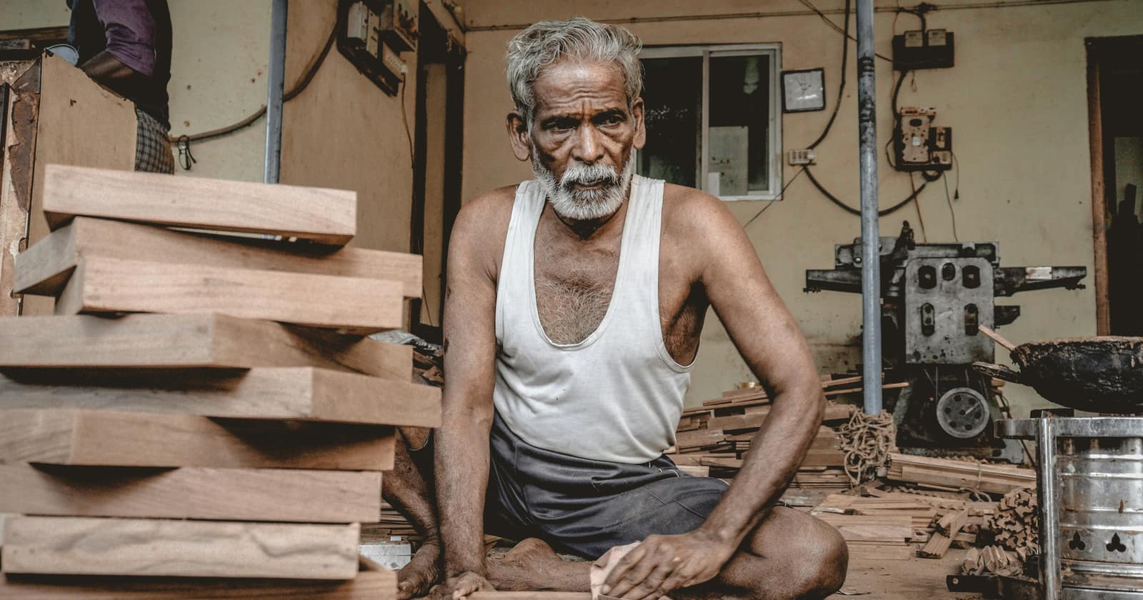 An elderly carpenter with a white beard seated on the floor of his workshop beside a stack of cut timber