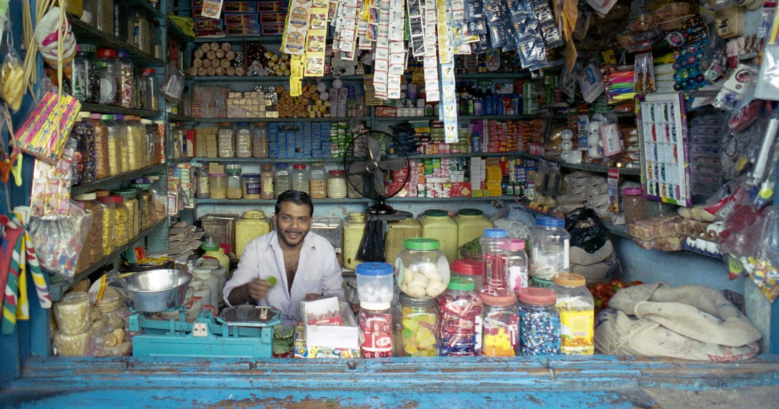 A smiling shopkeeper behind the counter of a small Indian store packed floor to ceiling with goods
