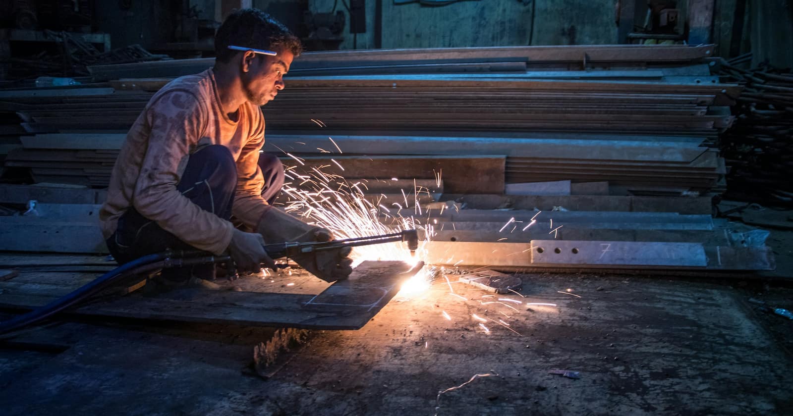 A worker crouched on a workshop floor cutting steel with a gas torch, sparks spraying across the ground