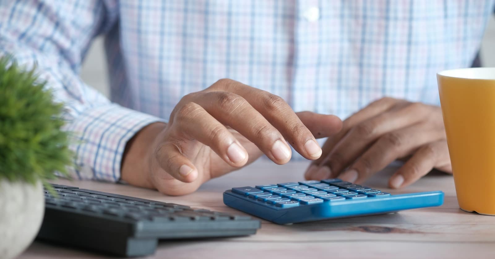 A person working a calculator at a desk beside a keyboard and a coffee mug