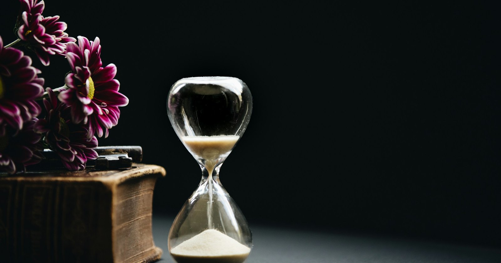 An hourglass with its sand running out beside flowers and an old book, on a dark table