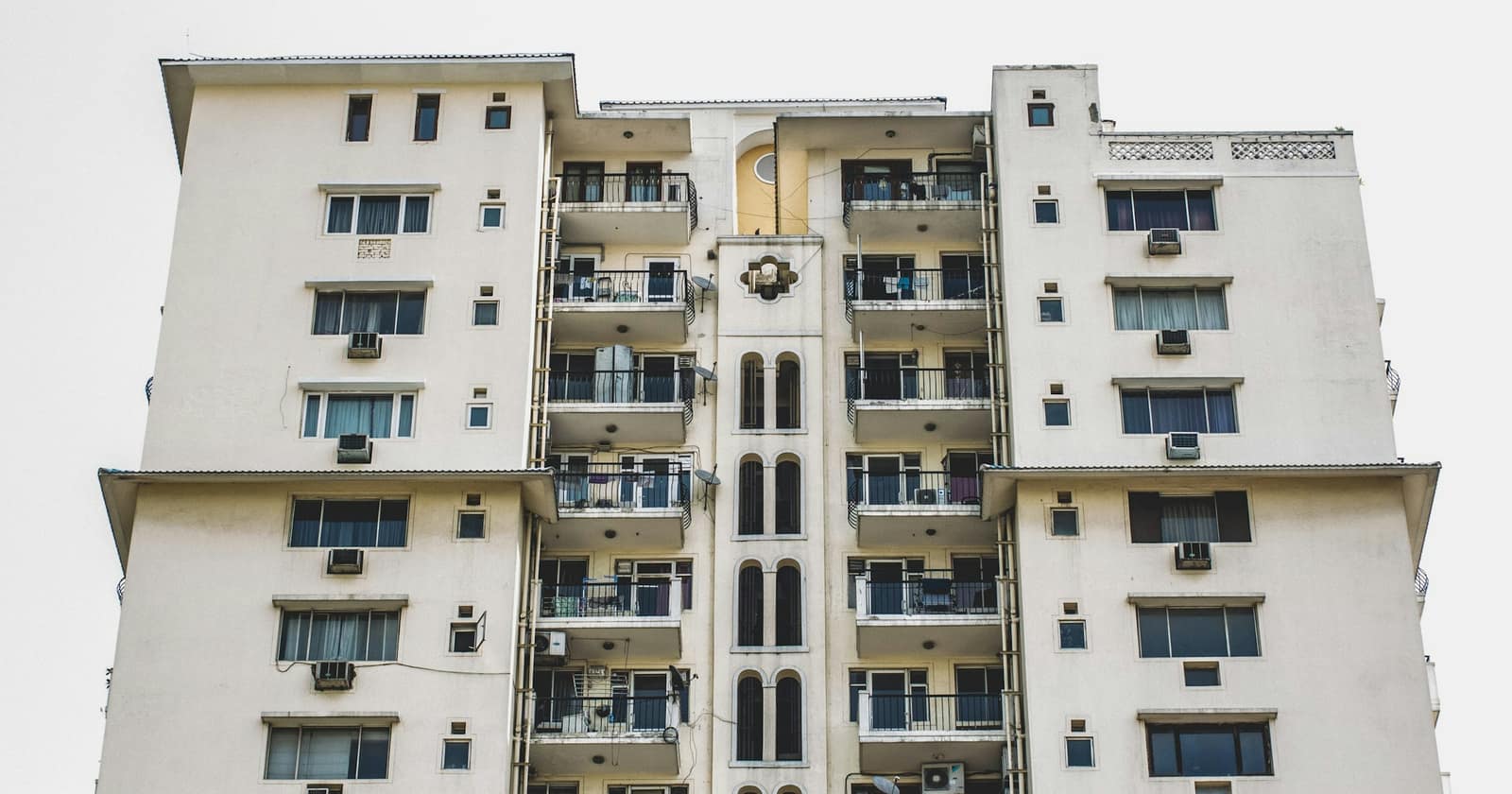 A white residential apartment block with balconies and air-conditioner units against a pale sky